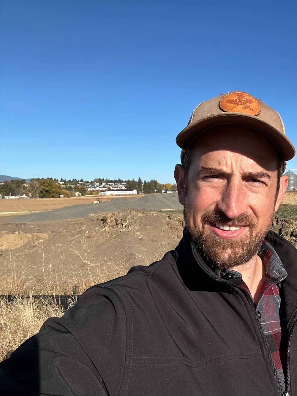 Charlton Haupt smiling in an open field, with grain silos and excavators behind him.
